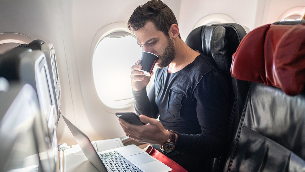 A man is sitting on the plane, drinking coffee and looking at his phone and laptop.