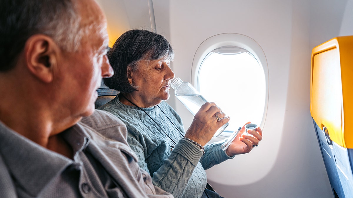Older couple seated on an airplane, with a woman drinking water by the window during a flight.