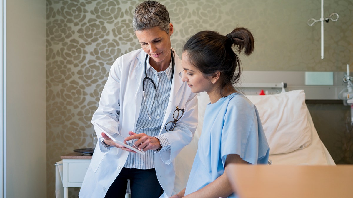 Doctor reviewing medical information with a young female patient sitting on a hospital bed in a private exam room.