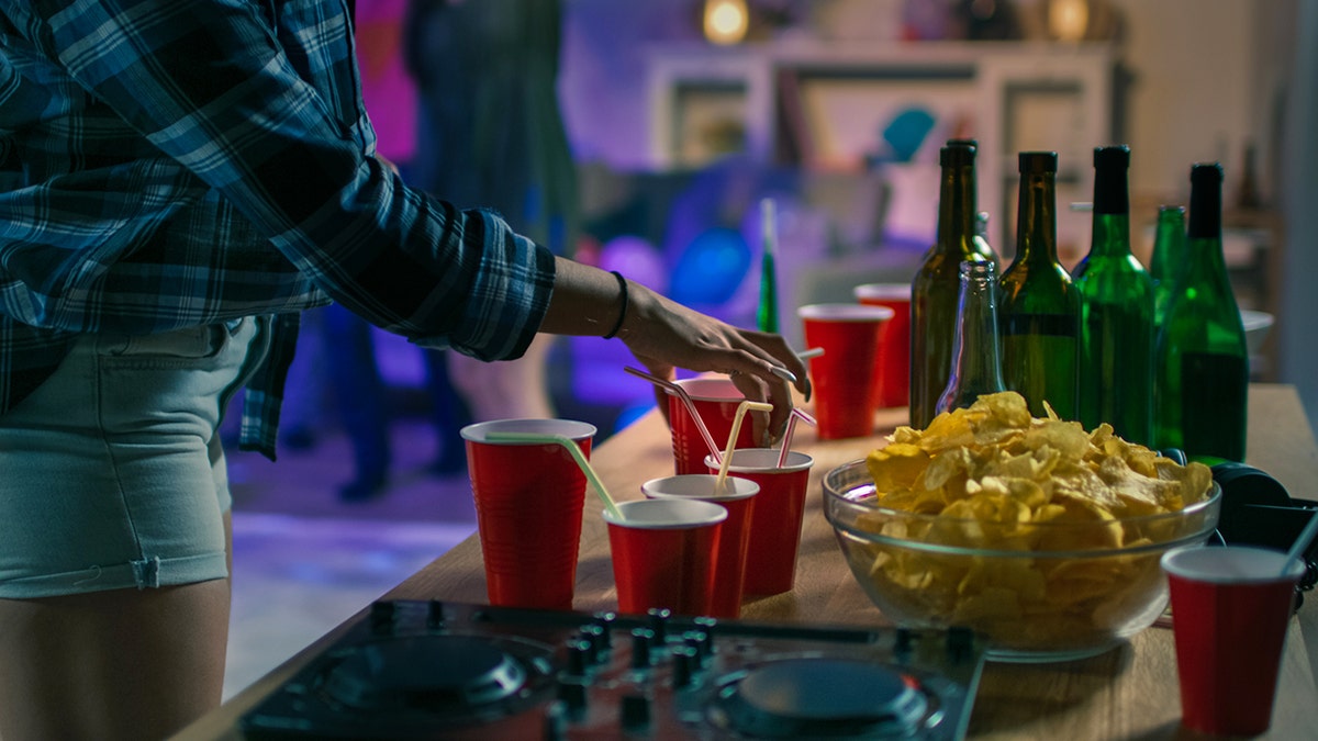 Party snack table with red cups, beer bottles, and a bowl of chips as a guest reaches for a drink