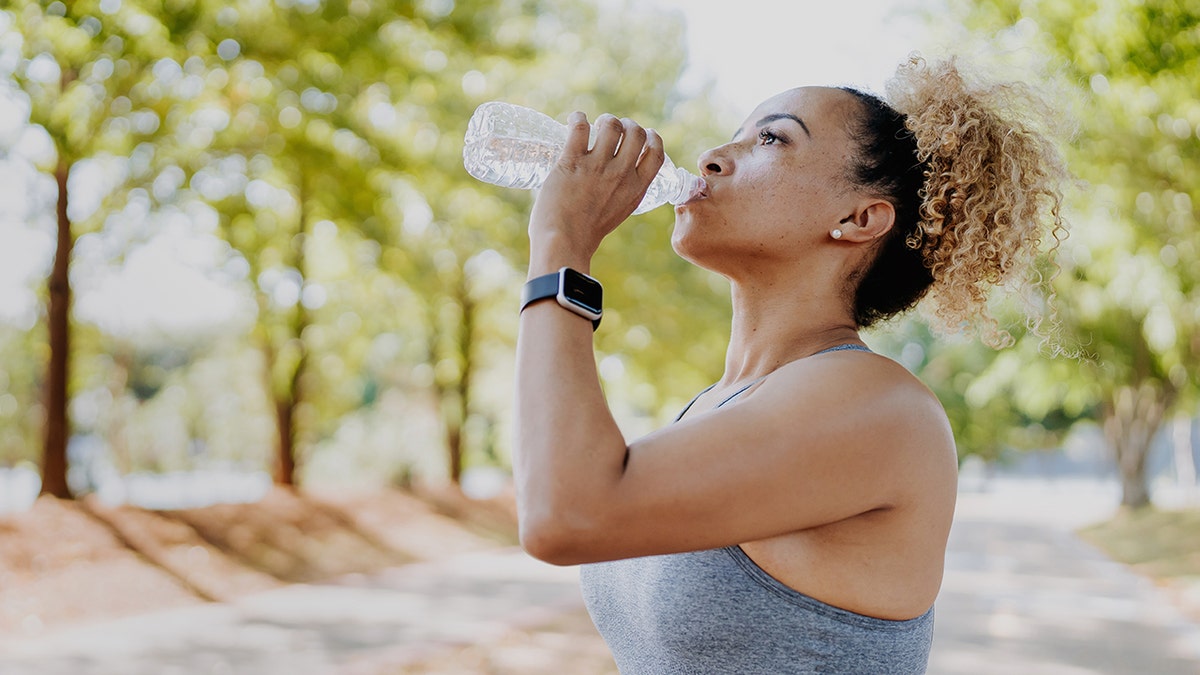 Photo of a woman drinking water from a plastic bottle in a green park surround by trees
