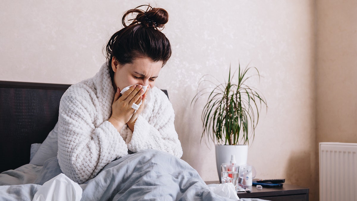 Woman sitting up in bed blowing her nose with a tissue, covered by a blanket and appearing sick at home.