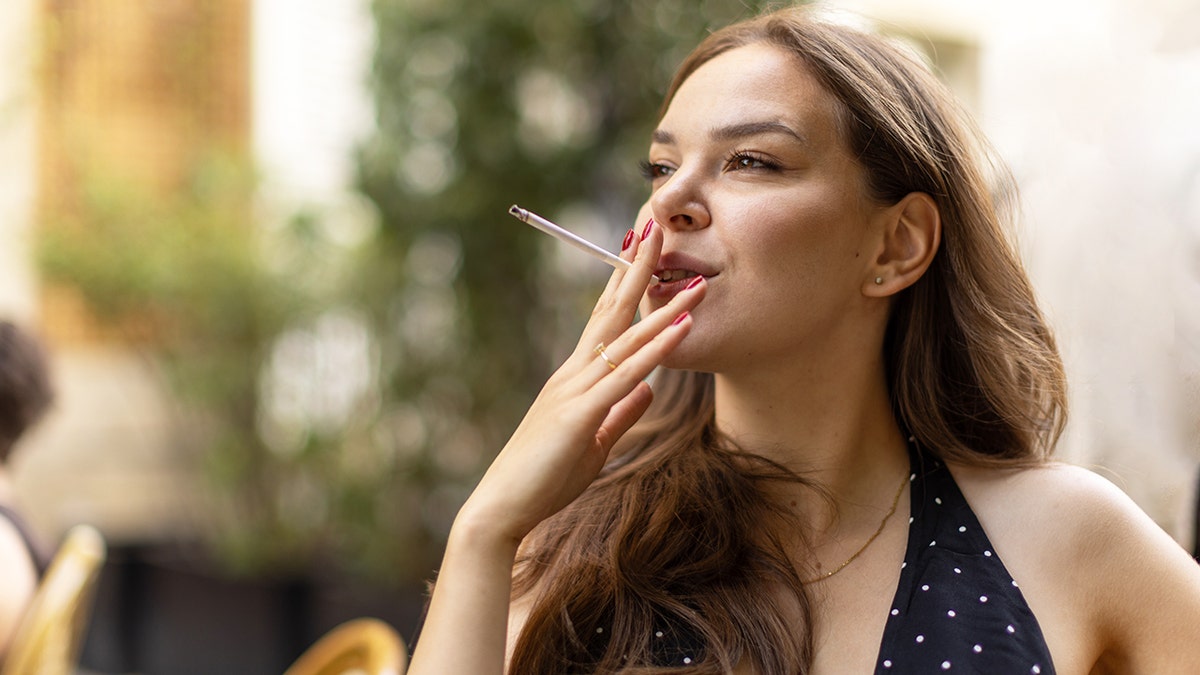 Young woman sitting outdoors, holding and smoking a cigarette.