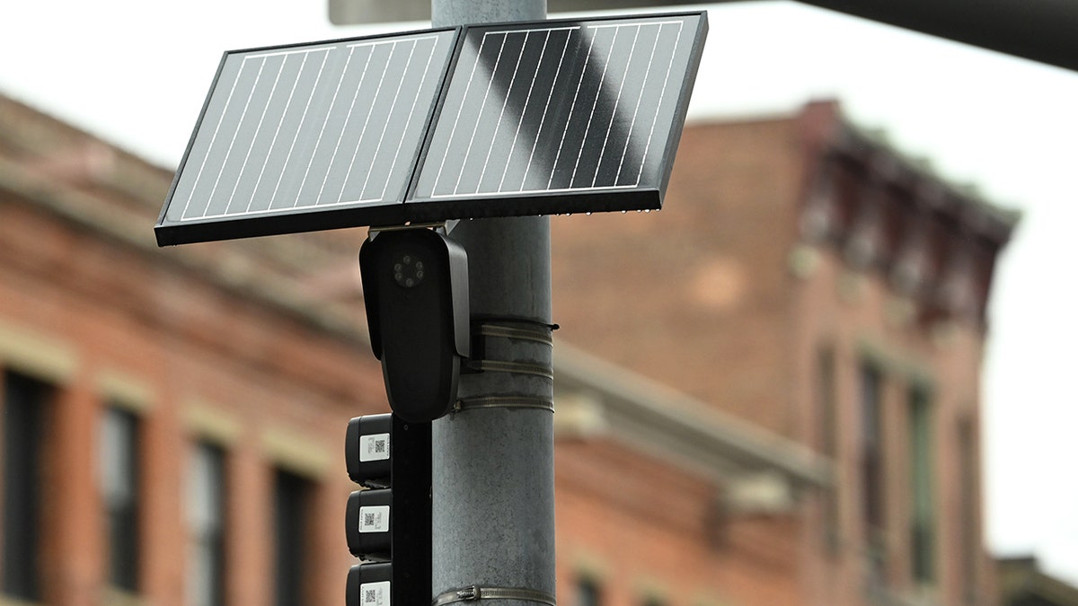 A Flock camera is secured to a light post with a solar panel above it.
