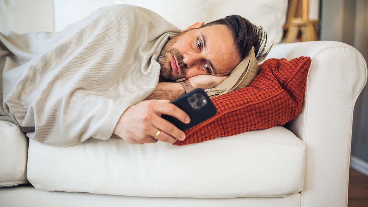 Man lying on a couch wrapped in a blanket while looking at his smartphone.