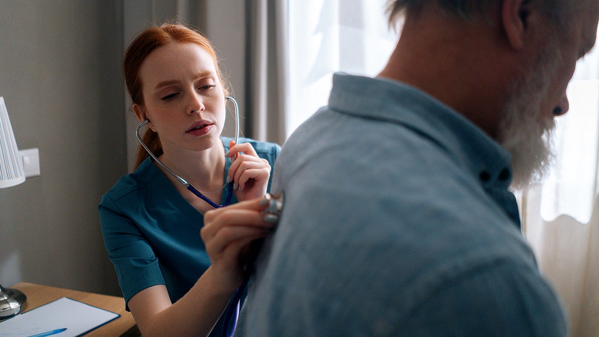 Nurse listening with stethoscope to back of mature adult man in hospital room.