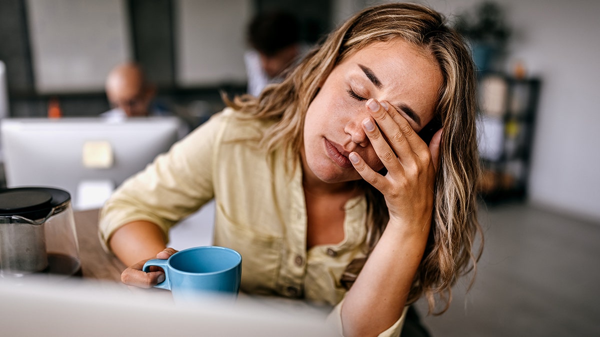 tired woman rubs eyes and holds coffee mug in office